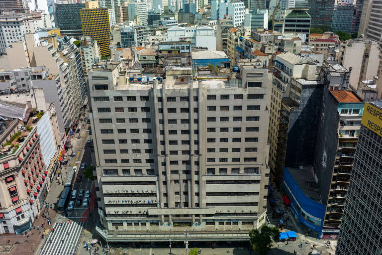 Imagem aérea mostra prédio ao centro. Ele tem cor bege e muitas janelas. É possível ver paredes pichadas. 
